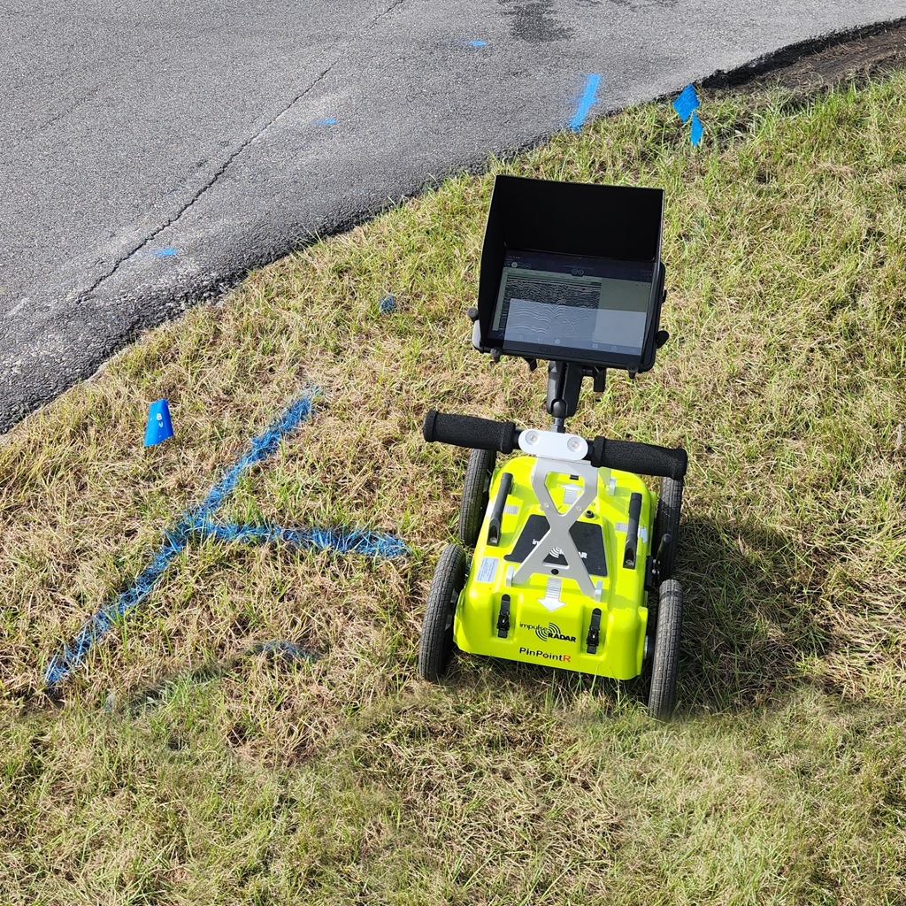 An ImpulseRadar PinPointR Ground Penetrating Radar GPR unit scanning the ground and revealing the location of a buried water line which has been marked with paint and flags on the grass.